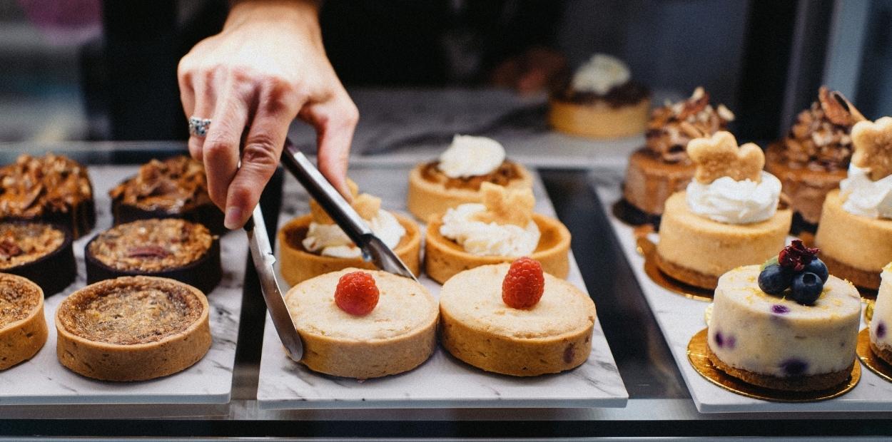 A display case full of pastries and mini cheesecakes decorated with fruit. A hand holding tongs is selecting a tart out of the case.